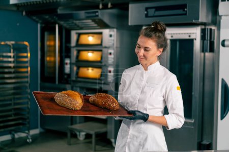 Téléchargez les photos : Chef souriante debout avec des pains fraîchement cuits dans la cuisine d'une boulangerie et regardant la caméra - en image libre de droit