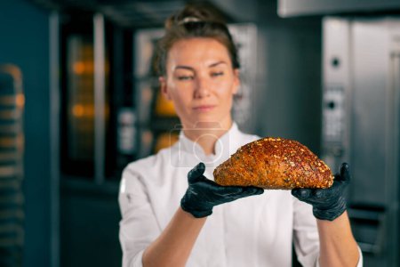 Photo for A smiling chef girl holds out freshly baked bread with crust and seeds to the camera while standing in the kitchen of a bakery - Royalty Free Image