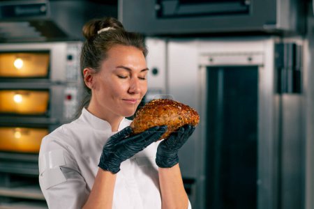 Téléchargez les photos : Portrait d'une fille dans une cuisine de boulangerie professionnelle qui inhale l'arôme du pain fraîchement cuit et sourit - en image libre de droit