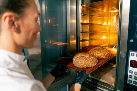 Téléchargez les photos : Une boulangère portant des gants sort une plaque de cuisson avec du pain chaud fraîchement cuit du four - en image libre de droit