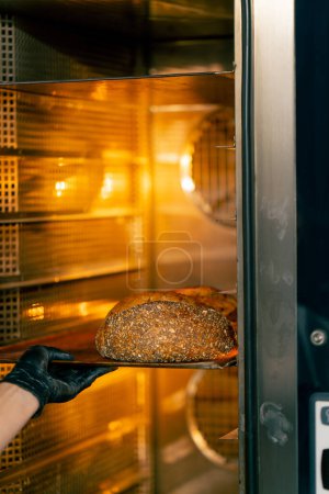 Photo for A female baker wearing gloves takes out a baking sheet with freshly baked hot bread from the oven - Royalty Free Image