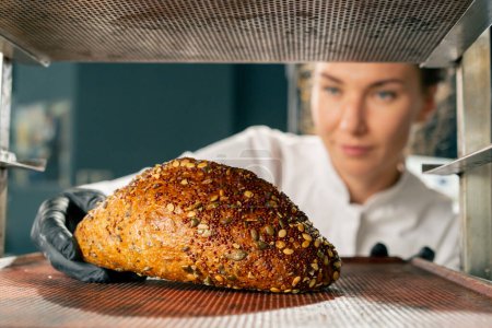 Foto de Una pastelera con guantes saca una bandeja para hornear con pan caliente recién horneado del horno - Imagen libre de derechos