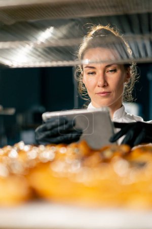 Photo for A focused bakery girl checks the number of baked puffs in the bakery against the delivery note in the clipboard - Royalty Free Image