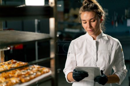 Photo for Portrait of a focused girl baker who checks the number of baked puffs in the bakery against a list in a tablet - Royalty Free Image