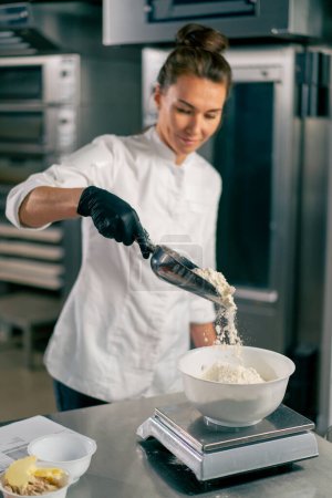 Photo for Female baker scoops wheat flour with a special spoon to knead dough for preparing fresh baked goods - Royalty Free Image