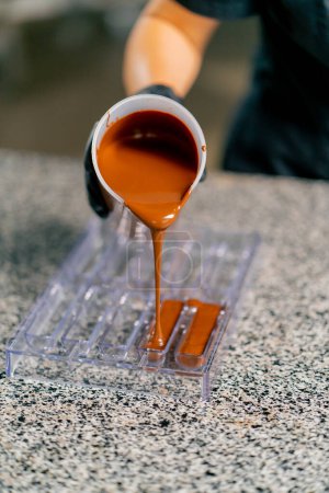 Téléchargez les photos : Le chocolat chaud fondu est versé dans un moule de chocolat transparent pour faire des bonbons naturels dans une confiserie - en image libre de droit