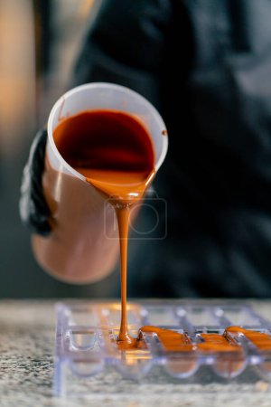 Foto de El chocolate caliente derretido se vierte en un molde de chocolate transparente para hacer caramelos naturales en una confitería - Imagen libre de derechos