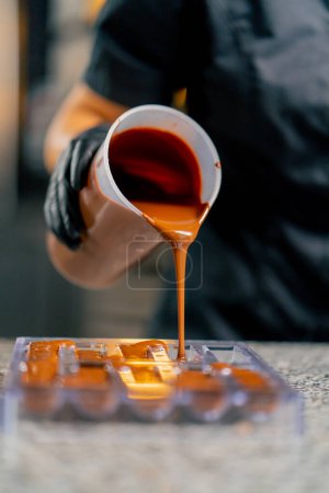 Téléchargez les photos : Le chocolat chaud fondu est versé dans un moule de chocolat transparent pour faire des bonbons naturels dans une confiserie - en image libre de droit