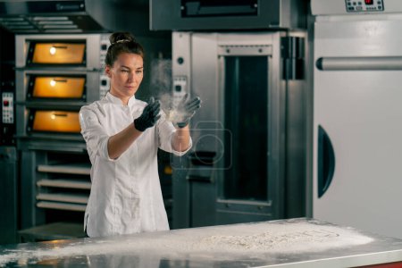 Téléchargez les photos : Une fille chef boulanger saupoudrer efficacement la farine sur une surface de cuisine pour préparer la pâte pour la cuisson des produits de boulangerie - en image libre de droit