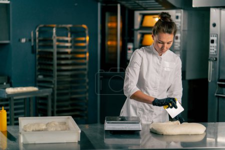 Photo for Female baker in uniform rolls dough into croissant shape on kitchen surface before baking in  oven - Royalty Free Image