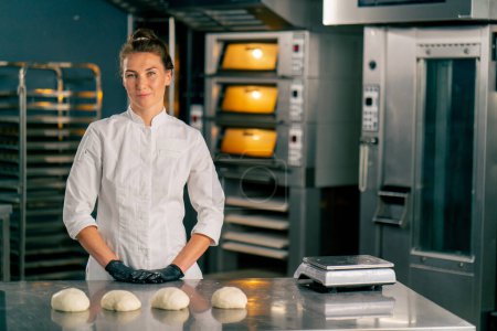 Téléchargez les photos : Femme boulanger se tient derrière une surface de cuisine professionnelle dans une boulangerie avec pâte préparée pour la cuisson et regarde la caméra - en image libre de droit