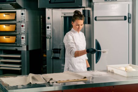 Téléchargez les photos : Une fille cuisinière dans une tunique se tient à la table de la cuisine et saupoudre la farine sur la pâte à travers un tamis - en image libre de droit