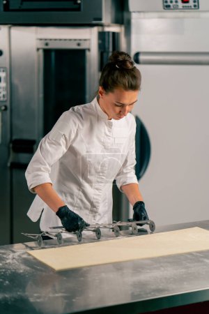 Foto de Mujer chef se para en la mesa de la cocina con un divisor de masa y corta la masa en partes iguales para hacer croissants - Imagen libre de derechos