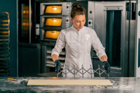 Photo for Woman chef stands at the kitchen table with a dough divider and cuts the dough into equal parts for making croissants - Royalty Free Image