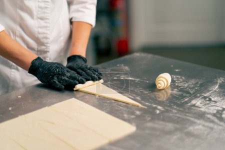 Foto de Primer plano de la mano de un chef en guantes rodando masa cortada cruda en formas de croissant para hornear - Imagen libre de derechos