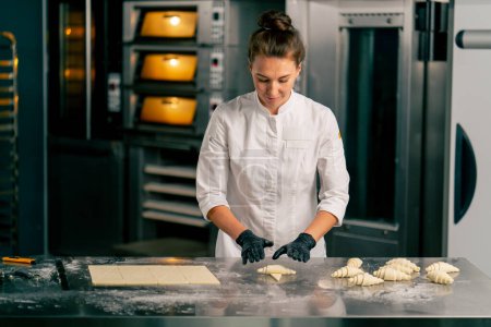 Foto de Una cocinera con chaqueta y guantes forma croissants de partes iguales de masa cruda para hornear por la mañana en una panadería - Imagen libre de derechos