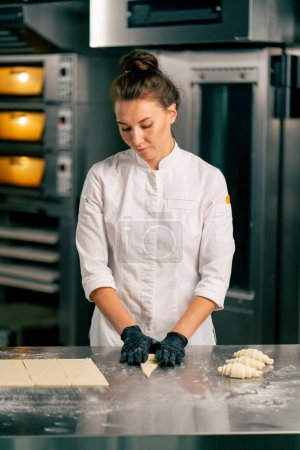 Foto de Una cocinera con chaqueta y guantes forma croissants de partes iguales de masa cruda para hornear por la mañana en una panadería - Imagen libre de derechos