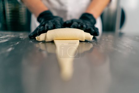 Foto de Primer plano de las manos enguantadas del chef dando forma y torciendo la masa cruda en forma de croissants para hornear - Imagen libre de derechos