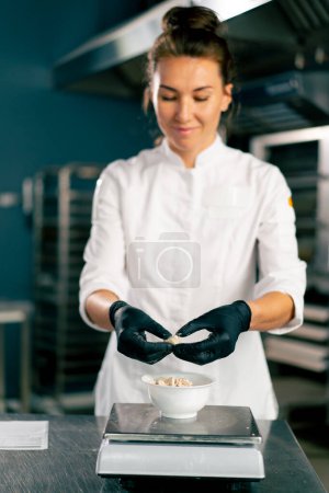 Photo for A female baker in a white chef's tunic kneads dough to prepare and bake bread in the bakery - Royalty Free Image