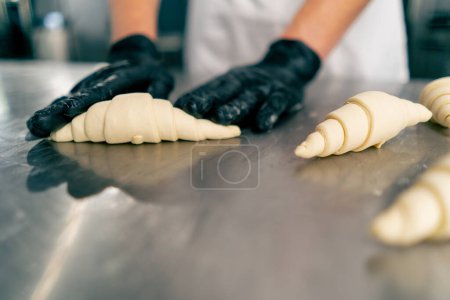 Téléchargez les photos : Gros plan des mains gantées du chef façonnant et tordant la pâte crue en forme de croissants pour la cuisson - en image libre de droit