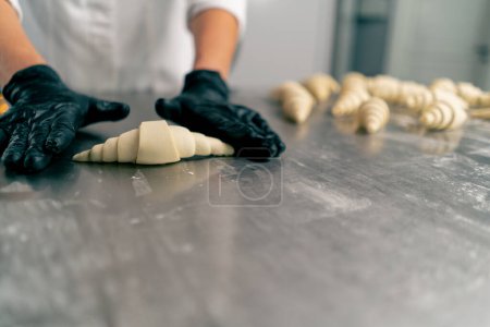 Foto de Primer plano de las manos enguantadas del chef dando forma y torciendo la masa cruda en forma de croissants para hornear - Imagen libre de derechos