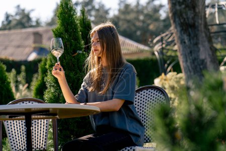 Téléchargez les photos : Vue latérale d'une fille adulte assise à une table dans le jardin avec un verre de vin et dégustation de la boisson - en image libre de droit