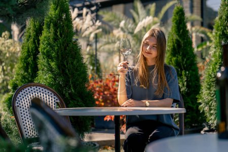 Foto de Una chica adulta se sienta en una mesa en el jardín con una copa de vino y prueba una bebida en el jardín en una bodega - Imagen libre de derechos