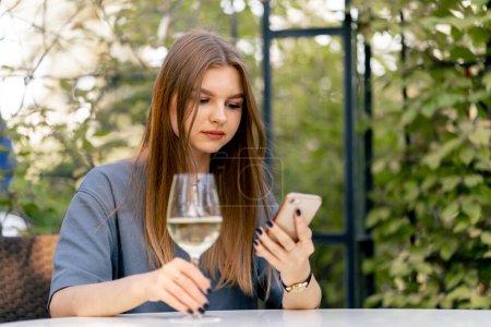 Téléchargez les photos : Triste jeune fille assise à une table dans le jardin avec un verre de vin et regardant le téléphone - en image libre de droit