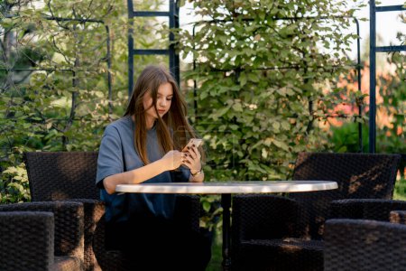 Téléchargez les photos : Une jeune fille triste s'assoit à une table dans le jardin et regarde soigneusement le téléphone dans ses mains - en image libre de droit