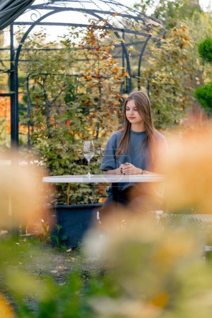 Téléchargez les photos : Une fille adulte s'assoit à une table dans le jardin avec un verre de vin et goûte une boisson dans le jardin dans une cave - en image libre de droit