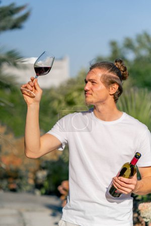 Téléchargez les photos : Charmant sommelier masculin lève un verre de vin naturel vieilli au soleil pour vérifier sa qualité et son goût - en image libre de droit