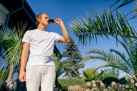 Foto de Un joven encantador se encuentra en la terraza de un restaurante cerca del jardín y toma un sorbo de delicioso vino. - Imagen libre de derechos