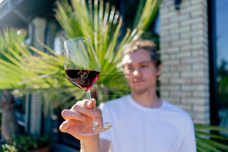 Foto de Un joven encantador sostiene una copa de vino a la cámara y examina cuidadosamente la bebida al sol - Imagen libre de derechos
