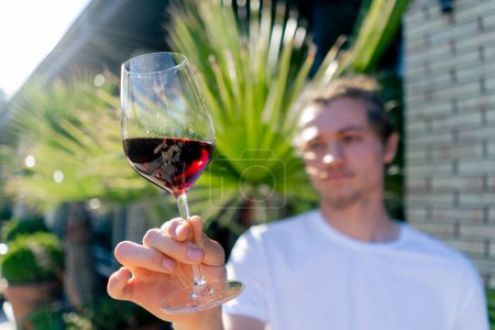 Téléchargez les photos : Un jeune homme charmant tend un verre de vin à la caméra et examine attentivement la boisson au soleil - en image libre de droit