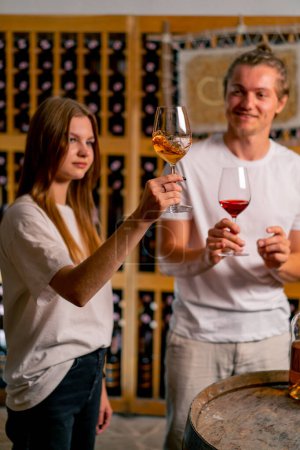 Foto de Una chica en una cata de vinos escucha atentamente a un sommelier masculino y prueba el vino de una hermosa copa en una bodega - Imagen libre de derechos