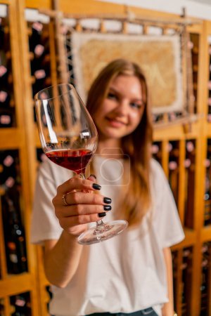 Photo for A girl waitress stands in a wine cellar holding a glass of wine in her hands and holds it out to the camera with a smile - Royalty Free Image