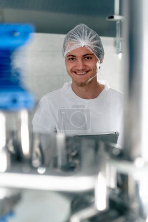 Foto de Retrato de un joven y sonriente tecnólogo en uniforme con una tableta cerca de los tanques en una producción cervecera - Imagen libre de derechos