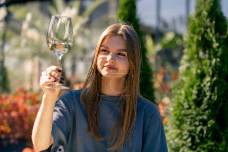 Téléchargez les photos : Une jeune belle fille tient un verre de vin dans sa main et l'examine soigneusement pour déterminer la qualité de la boisson - en image libre de droit
