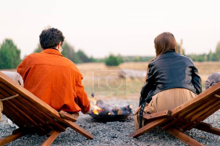 Foto de Volver tiro de un joven hombre y una mujer sentados en la naturaleza por el fuego relajarse y asar malvaviscos - Imagen libre de derechos