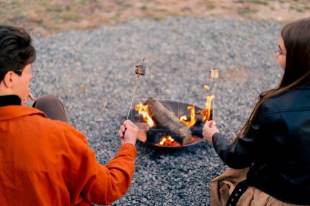 Téléchargez les photos : Plan arrière d'un jeune homme et d'une jeune femme assis dans la nature par un feu relaxant et rôtissant des guimauves - en image libre de droit