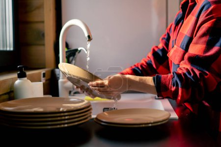 Foto de Una chica con una camisa a cuadros lava a fondo los platos con espuma en la cocina de una casa de madera - Imagen libre de derechos