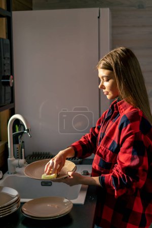 Foto de Una chica con una camisa a cuadros lava a fondo los platos con espuma en la cocina de una casa de madera - Imagen libre de derechos