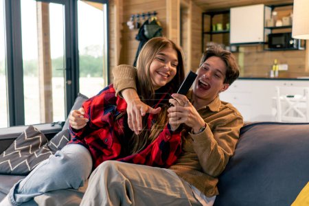 Téléchargez les photos : Jeune couple amoureux s'amuser, rire et se battre pour la télécommande de la télévision tout en étant assis sur le canapé - en image libre de droit