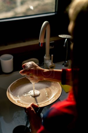 Téléchargez les photos : Gros plan de la main d'une femme avec une éponge en mousse lave-vaisselle sous la pression de l'eau dans une cuisine - en image libre de droit