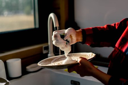 Foto de Primer plano de la mano de una mujer con una esponja de espuma lavar platos bajo la presión del agua en una cocina - Imagen libre de derechos