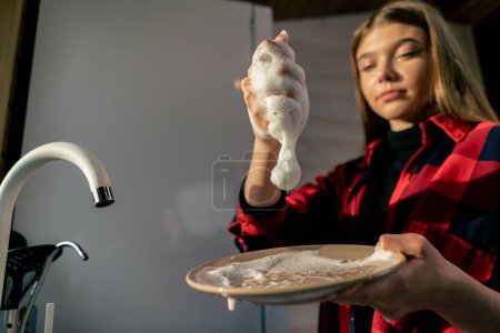 Foto de Una joven en una camisa esponja con espuma lava platos bajo la presión del agua en la cocina de la casa - Imagen libre de derechos