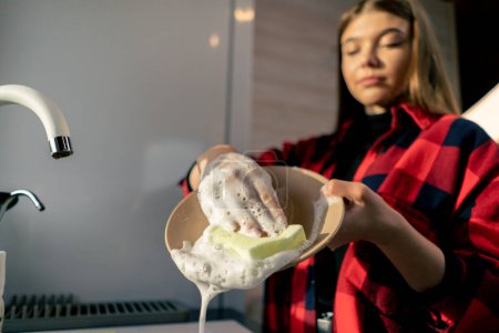 Téléchargez les photos : Une jeune fille dans une éponge chemise avec mousse lave les assiettes sous la pression de l'eau dans la cuisine de la maison - en image libre de droit