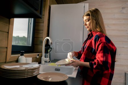 Foto de Una joven en una camisa esponja con espuma lava platos bajo la presión del agua en la cocina de la casa - Imagen libre de derechos