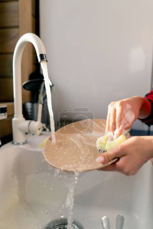 Foto de Primer plano de la mano de una mujer con una esponja de espuma lavar platos bajo la presión del agua en la cocina - Imagen libre de derechos