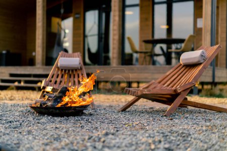 Foto de Un fuego está ardiendo al aire libre cerca de una acogedora casa de madera con terraza en un sitio de glamping en la estepa - Imagen libre de derechos